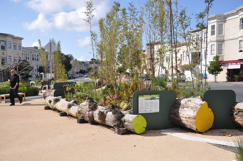A daytime view of Guerrero Plaza featuring a row of large wooden logs and green informational panels separating a sandy pedestrian path from a landscaped area with young trees. San Francisco-style residential buildings and San Jose Avenue are visible in the background.